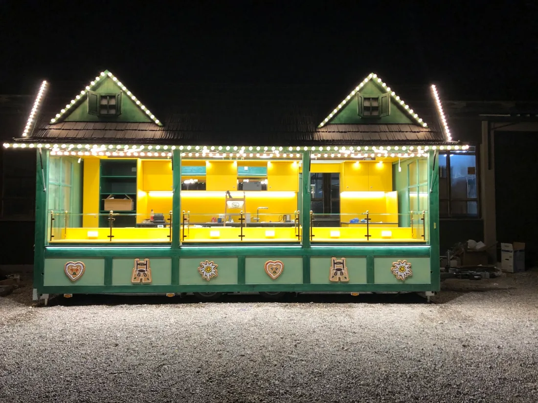 Beleuchteter Verkaufswagen im Almhaus-Stil bei Nacht — grün mit Giebeln, Lichterketten und Lebkuchenherzen